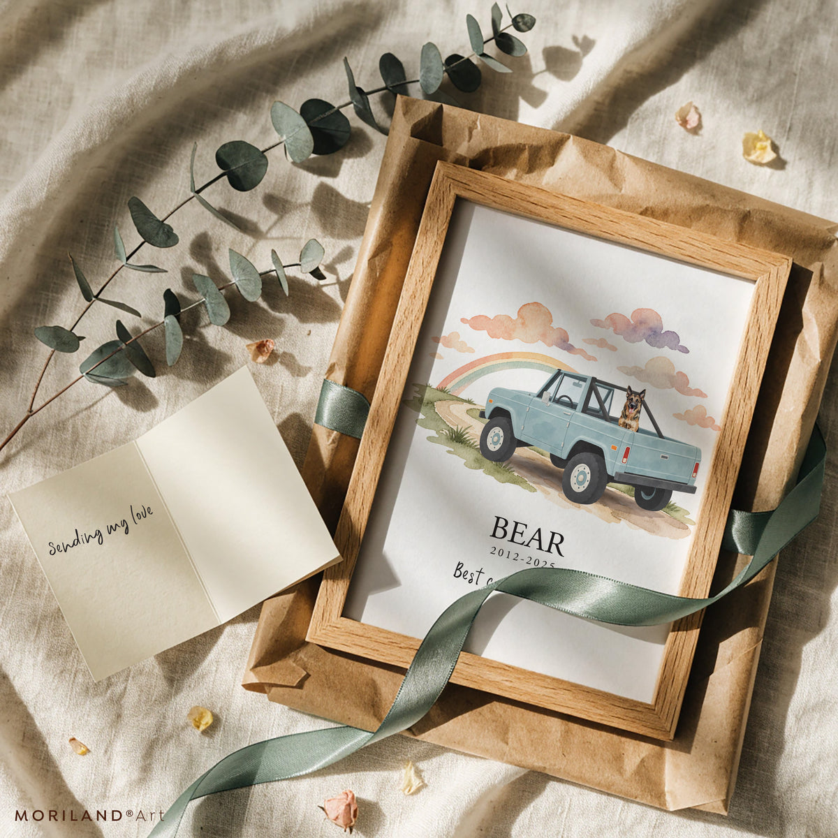 Pet remembrance gift: framed watercolor print of a German shepherd in a vintage open-top 4x4 with the name Bear, wrapped in kraft paper with a green ribbon, eucalyptus and a sympathy card