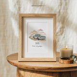 Small framed dog memorial print of a golden retriever in a pale blue vintage pickup driving toward a rainbow, displayed on a round wooden side table next to a lit candle