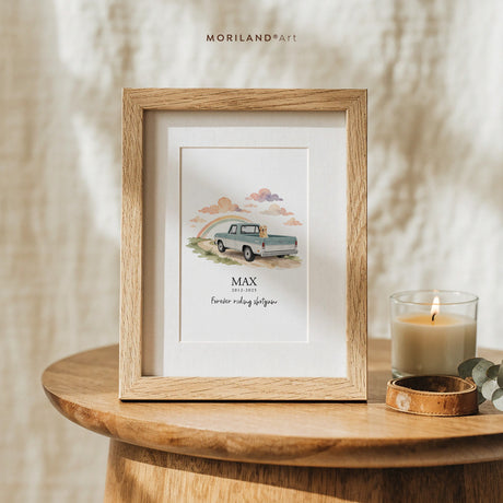 Small framed dog memorial print of a golden retriever in a pale blue vintage pickup driving toward a rainbow, displayed on a round wooden side table next to a lit candle