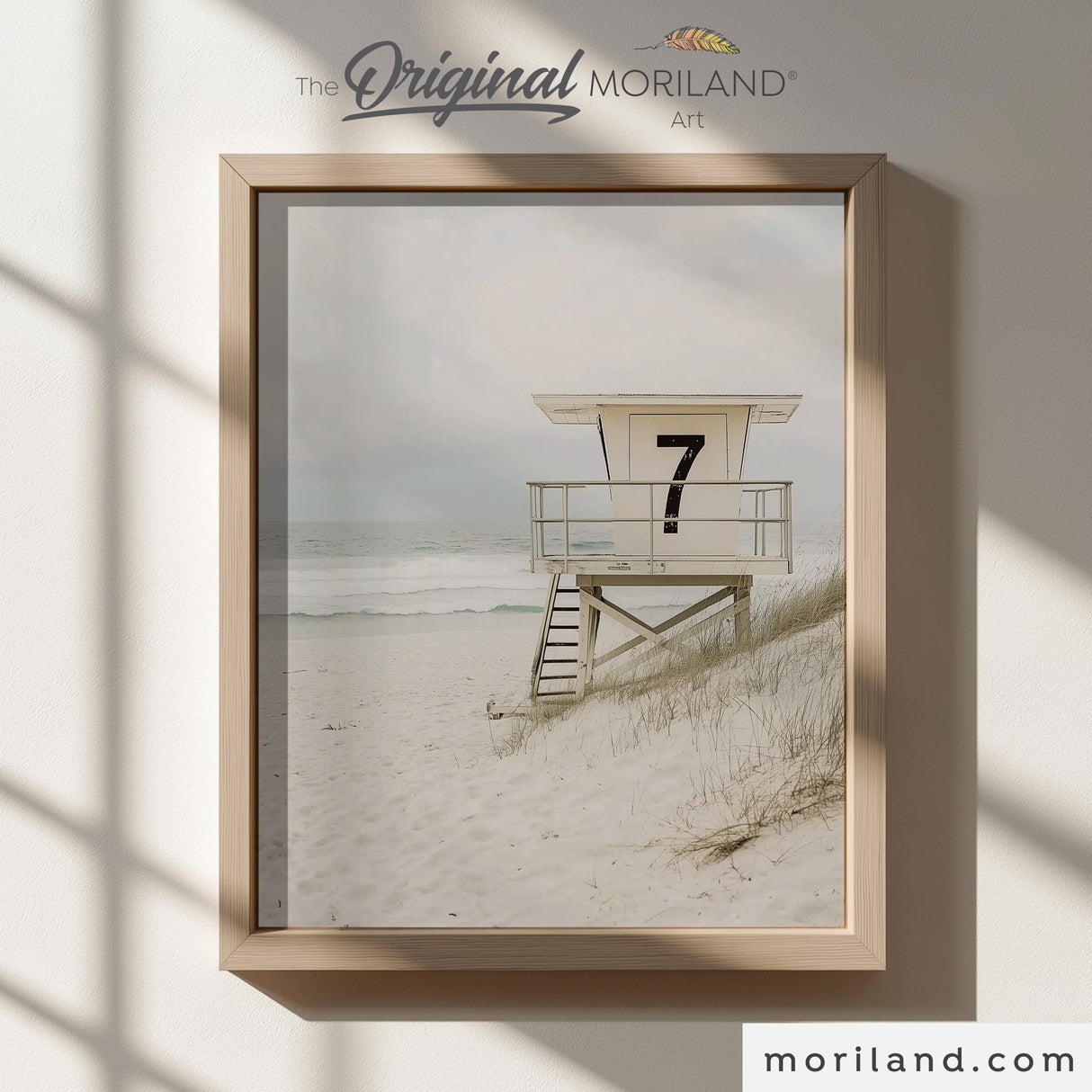 Modern heritage coastal style photographic print of a vintage white wooden lifeguard tower stand with the number 7 on sandy dunes by the ocean, displayed in a light wood frame for a beach nursery or coastal playroom by MORILAND.