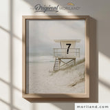 Modern heritage coastal style photographic print of a vintage white wooden lifeguard tower stand with the number 7 on sandy dunes by the ocean, displayed in a light wood frame for a beach nursery or coastal playroom by MORILAND.